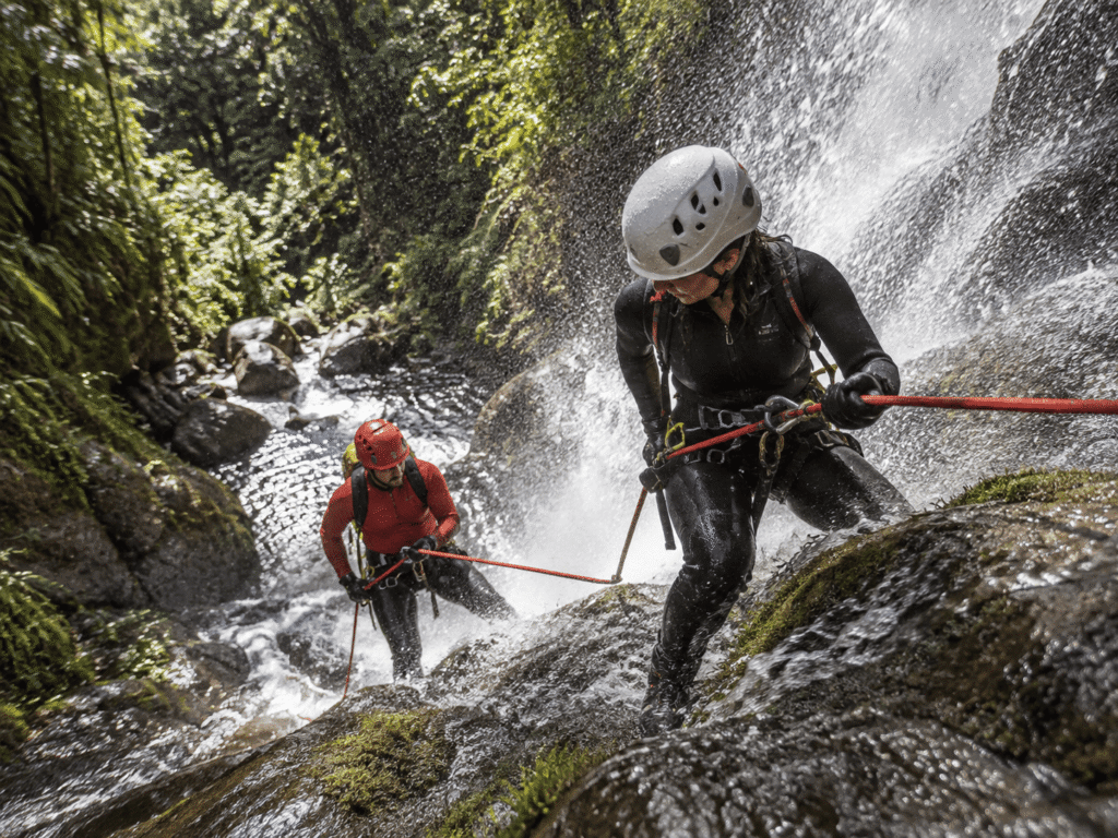 Praticantes com capacetes e equipamentos de corda em uma cachoeira cercada por vegetação.